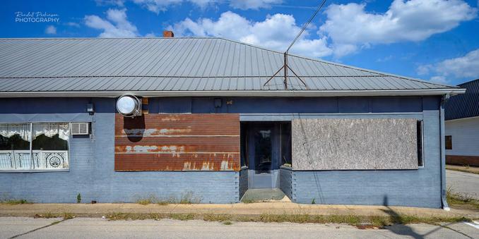 A one story brick building painted grey has one recessed entry door, one window covered with plywood, and a second window covered in rusting corrugated metal.  A large vent fan and a window AC unit hang off the side of the building near a third window.