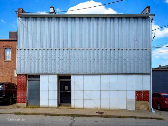 A two story brick building with one glass and metal entrance door and a second boarded up door is covered by 2' square white panels or tile on the bottom portion a two rows of different texture silver metal panels above the door height.  A portion of the brick building is visiting at the bottom left and right corners of the building.