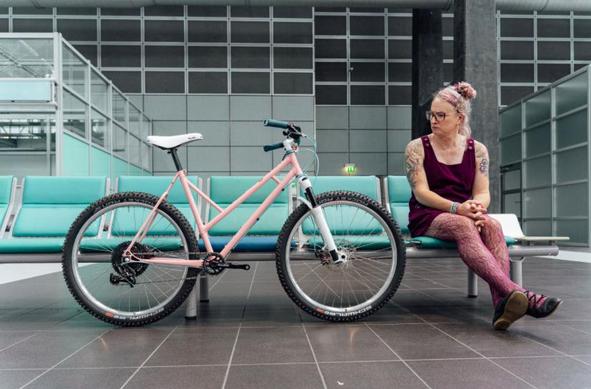 Elodie sits on a row of bright teal seats next to a pink mountain bike
She is wearing a burgundy pinafore dress and matching patterned tights