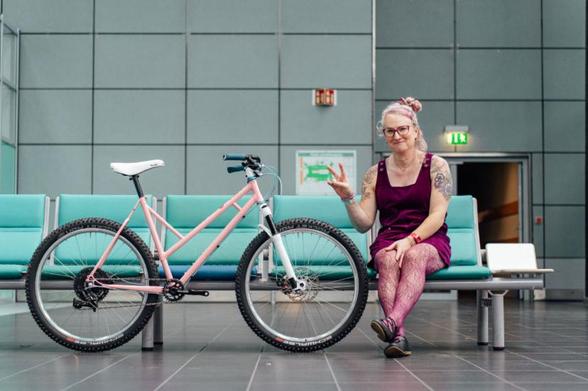 Elodie sits on a row of bright teal seats next to a pink mountain bike
She is wearing a burgundy pinafore dress and matching patterned tights
