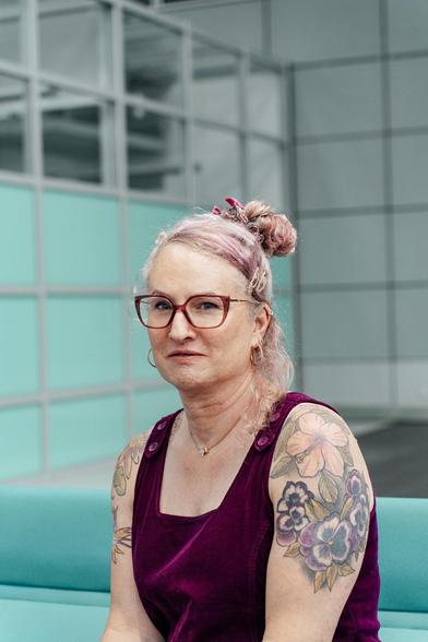 Elodie is looking into the camera
She is wearing a burgundy pinafore dress and pink glasses
Flower tattoos are visible across her shoulders and upper arms