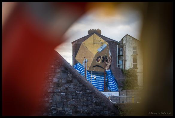 A large-scale street mural titled "What is Home" viewed through the circular opening of a red and white life buoy, depicting a figure in a blue and white striped shirt reaching up towards geometric shapes, painted on the gable end of a building with stone walls visible in the foreground.