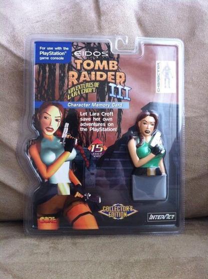 Blister pack showing a Tomb Raider III Collector’s Edition PlayStation memory card. Packaging features artwork of Lara Croft with pistols, plus a 3D bust-shaped memory card in green top and brown shorts. Text reads “15 blocks of memory.”
