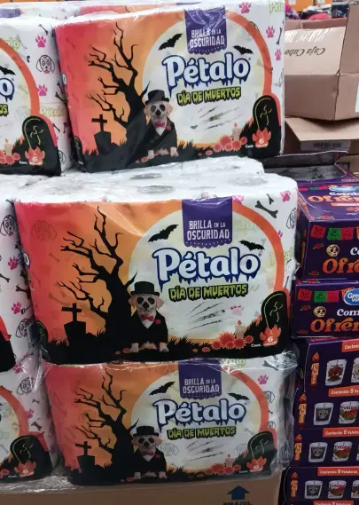 A stack of three packs of Pétalo toilet paper.

The packs say "DÍA DE MUERTOS" and have an image of a skull-faced dog in a jacket, top hat and bow tie, a dead tree, gravestones, cempazúchitl marigolds, and bats flying across the setting sun.

The packs are also marked "BRILLA EN LA OBSCURIDAD" (glows in the dark).

Seen in a branch of Bodega Aurrera supermarket.