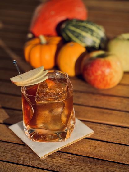 a sunlit, amber brown cocktail with apple slice fan garnish and big rock of clear ice in a warped hand-blown glass sitting on a white coaster on a wooden table with pumpkins and apples in the background