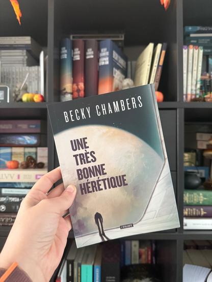 Livre pris en photo devant une bibliothèque. 
La couverture d’Une très bonne hérétique montre une créature bipède sur une surface de vaisseau/station spatiale avec en face une planète.