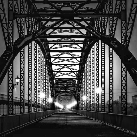 Frontalaufnahme der Alten Elbbrücke Hamburg Harburg zur Blauen Stunde. Die Brücke ist von einer Reihe alten Lampen, die an der Stahlkonstruktion befestigt sind, beleuchtet. Es handelt sich um eine Schwarz Weiss Aufnahme.