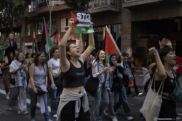 Manifestación. una joven grita y alza un cartel que dice "STOP GENOCIDIO" junto a un grupo que porta banderas palestinas y aplaude. Ambiente de protesta y solidaridad.