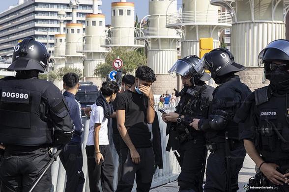 Grupo de policías antidisturbios con equipamiento completo identifica a un grupo de menores en la calle. Uno de los jóvenes se cubre el rostro con la mano. Fondo urbano con edificios y semáforos.