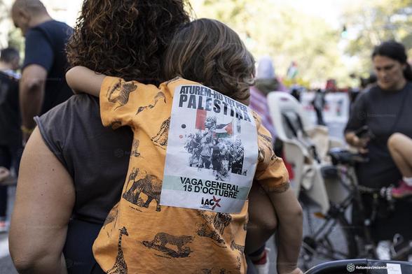 Madre con niñe pequeño a la espalda en una manifestación, con una camiseta amarilla que lleva un cartel adhesivo en la espalda: "Aturem-ho tot per Palestina. Vaga general 15 d’octubre. iAC". Al fondo, más gente y carteles, en un ambiente de protesta.
