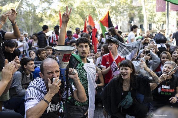 Manifestación multitudinaria: un manifestante con megáfono y pañuelo palestino grita consignas, detrás otro hace el gesto de victoria, mientras la multitud aplaude y muestra apoyo. Ambiente de energía y solidaridad.