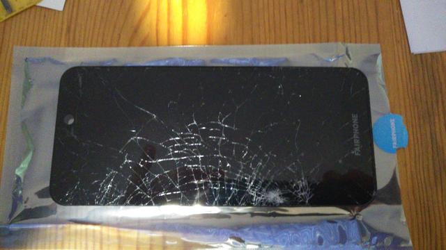 Image of a very shattered fairphine mobile phone screen resting on a grey shiny bag with a blue circular fairphone sticker on the right hand side. The phone is in a landscape position on a wooden table.