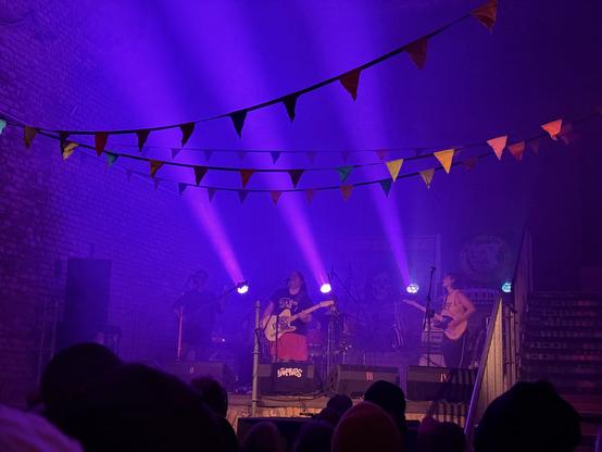 A live music performance with musicians on stage illuminated by vibrant purple lights. Colorful bunting hangs overhead, adding to the festive atmosphere. The audience is visible in the foreground, watching the show.