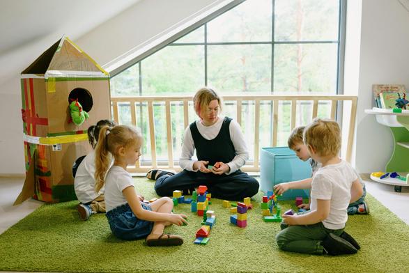 Das Bild zeigt eine Gruppe von Kindern und einer Erwachsenen in einem hellen Raum mit großen Fenstern, die viel natürliches Licht hereinlassen. Die Kinder sitzen auf einem grünen Teppich und bauen mit bunten Bausteinen. Eine Frau, wahrscheinlich eine Lehrerin oder Betreuerin, sitzt in der Mitte und hilft den Kindern bei ihrem Bauwerk. Einige Kinder bauen mit den Steinen, während andere zuschauen. Im Hintergrund ist ein aus Karton gebauter Vogelhaus zu sehen, der mit bunten Bändern verziert ist. Der Raum ist freundlich und einladend, mit einem Bücherregal und Spielzeug im Hintergrund.