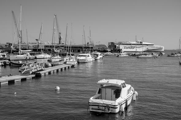 A black and white photograph of the harbour at Guernsey. A sunny day with a blue sky. In the foreground is a small cabin cruiser. On the left of frame is a jetty and beyond are many moored craft. In the background are cranes with jibs raised and at the upper right of frame is the Condor Ferry, leaving the harbour.