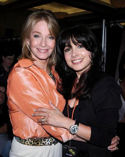 A 2011 photo of Deidre Hall and Judy Strangis taken at the Hollywood Autograph Show in Burbank, California. Deidre, known for her role as Marlena Evans on Days of Our Lives, wears a shiny peach blouse tucked into white pants with a leopard-print belt. Her straight, shoulder-length blonde hair frames a warm smile. Judy, known for her role as DynaGirl in ElectraWoman and DynaGirl, wears a black blouse or jacket over a black top, with long dark brown or black hair cascading past her shoulders. They are hugging and smiling directly at the camera. The background is dimly lit, suggesting an indoor convention setting. The image radiates warmth, nostalgia, and the enduring charm of two television icons reconnecting.