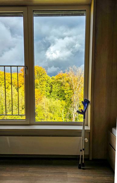 Ein Paar blaue Unterarmgehstützen steht neben einem großen Fenster, durch das herbstlich bunt gefärbte Bäume und ein dramatisch bewölkter Himmel zu sehen sind.