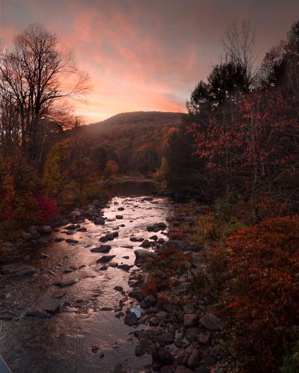 Looking over the middle of a dark river at dawn. There are rocks in the river and trees on both sides. It’s an autumnal scene with muted reds and yellows on the trees and the sky is pink with wispy clouds
