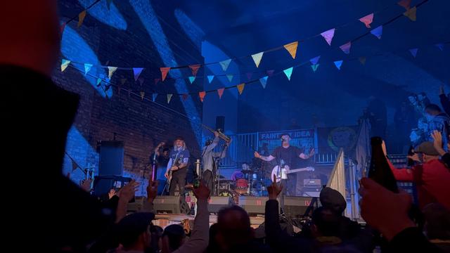 A live music performance featuring a band on stage with colorful bunting overhead. Audience members are cheering and raising their hands. The venue has a brick wall backdrop and various instruments on stage.