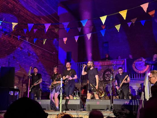 A live band performs on stage in a dimly lit venue, decorated with colorful bunting. Musicians play various instruments, including guitars and brass. Audience members are visible in the foreground, enjoying the performance. Flags and banners decorate the background.