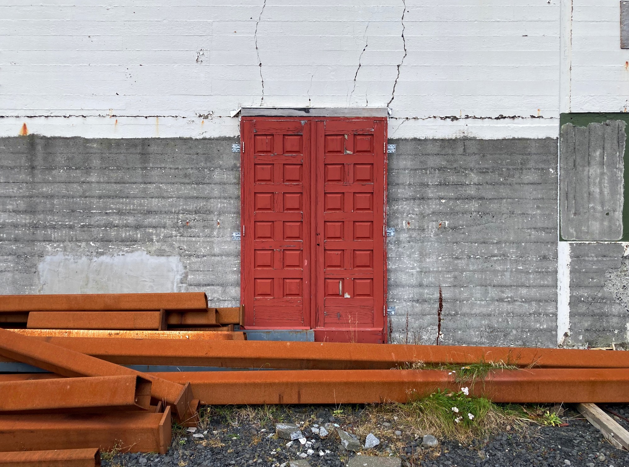 An old red door with cracked paint against a concrete wall with rusty beams lying on the ground.