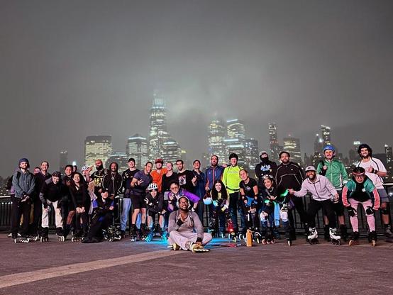 Group photo of 30+ Tuesday Night Skaters in the Jersey shoreline with the misty lights of lower Manhattan behind them.