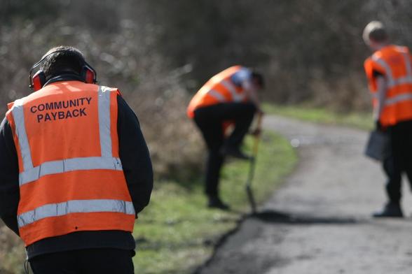 Three people in orange hi‑vis vests on a Community Payback scheme. One, with “Community Payback” written on his back and wearing ear protectors, faces away from the camera. Another digs at the side of a footpath while a probation worker looks on holding a clipboard.