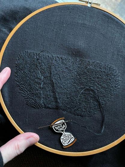 Lia’s fingers show a wooden embroidery hoop with a trapezoid of black on black whipped backstitch that looks a bit like flames, but if you’ve been following this piece you’ll know it’s pubic hair. It’s definitely pubic hair. The trapezoid is 14cm wide along the top, 7cm wide along the bottom, and 8cm tall. There’s white chalk markings within the hairs. An hourglass-shaped needle minder that reads “this is taking forever” is attached to the cloth.