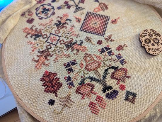 photo of a traditional cross stitch sampler featuring autumn colors and motifs of leaves and flowers. you can also see my needle minder which is a calavera-styled skull.