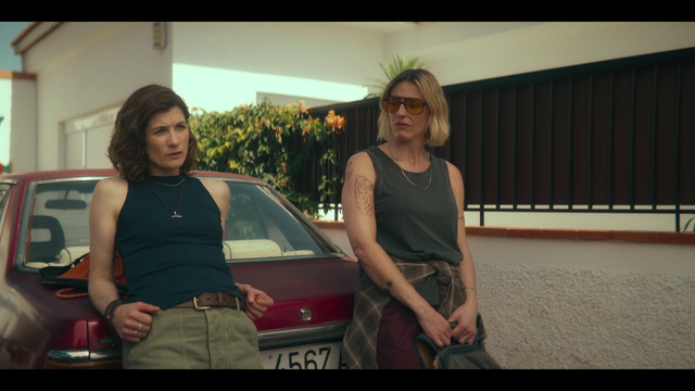 A screencap of Frauds, showing Jodie Whittaker's character Sam and Suranne Jones' character Bert leaning against the back of a car. Sam is on the left, with shoulder-long brunette hair, wearing a black tank and green shorts. Bert is on the right, with shorter blonde hair that shows her brown roots coming in, orange sunglasses, a green tank and red pants. She also has a longsleeve folded around her hips and holds a bag. You can see bits and pieces of her tattoos on her arms. In the back, there is a white house and a brown balcony fence with flowers hanging over it.
