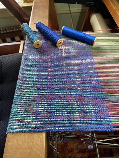 Photo of weaving in progress on the loom from a side angle.  The design is a multi-color echo twill, the colors bleed into each other on the diagonal.  There are three spools of different shades of blue weft on the far end that correspond to the section of the weft in which they were woven.  On the left is a teal, the middle is royal blue and the right is a brighter royal blue.  You can see the difference in shades in the weaving.