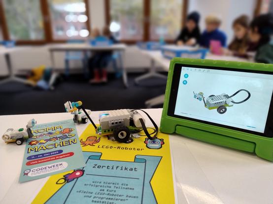 Auf einem Tisch liegt ein Flyer zur Code Week und eine Teilnehmerurkunde. Darauf steht ein aus Lego gebauter Roboter. Rechts daneben ein Tablet mit der Bauanleitung.
Unscharf im Hintergrund sitzen Kinder an Tischen und bauen und programmieren diesen Roboter nach.