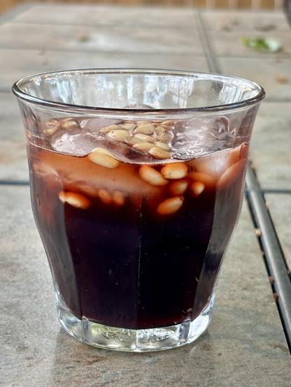 A clear glass filled with jallab, a dark reddish-brown Middle Eastern drink made from date syrup, topped with ice cubes and pale pine nuts floating on the surface. The glass sits on an outdoor table.