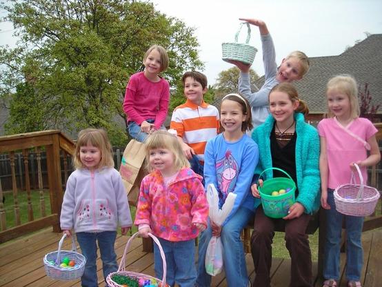 A group of eight smiling children are gathered on a wooden deck outdoors during what appears to be an Easter egg hunt. The kids are holding colorful baskets filled with plastic eggs and wearing casual spring clothes, including jackets, jeans, and bright tops. Some are sitting while others stand behind them, with a boy in the back playfully lifting an empty basket over his head. A large tree and suburban houses are visible in the background under a cloudy sky, giving the photo a cheerful, candid atmosphere of a family or neighborhood celebration.
