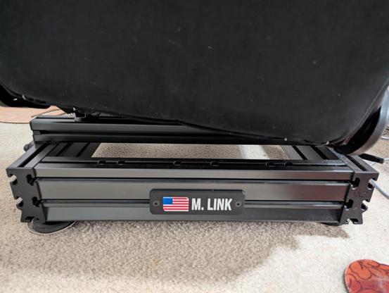 A close-up photograph of the base of a black **racing simulator seat**. The seat itself, which is also black, is only partially visible at the top of the frame. The base is constructed from black **aluminum extrusion profiles**, forming a sturdy, rectangular frame on the floor.

A small, rectangular, dark nameplate is attached to the front of the base. It features the **American flag** on the left and the white text "**M. LINK**" on the right. The entire structure rests on carpet, and the dark profile ends show a pattern of multiple bolt holes.