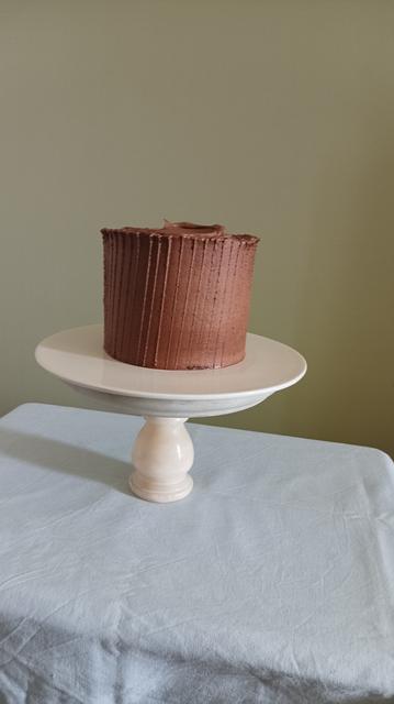 A tall chocolate cake with vertical line design running throughout its side kept on a white ceramic plate, that, in turn, is kept on a heavy curvy white marble cake stand. 

The whole thing rests on a table covered in baby blue table clothes (slightly creased), with a sage green wall in the background.