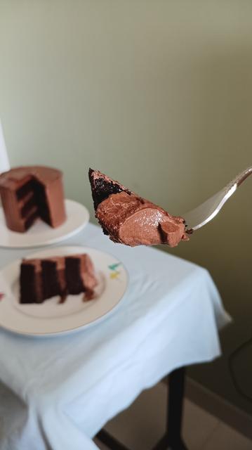 A fork holding a small portion of a chocolate cake (dark brown) with chocolate frosting (light brown) is angled towards the camera. 

In the background you can see a small table covered in baby blue table cloth. 

On the table is one cake plate with a blurry chocolate cake, out of which a slice has been taken out. Next to this is a white dinner plate with some blue green design right at the edge. There is a slice of chocolate cake kept on its side on this plate. 

Sage green wall in the background.