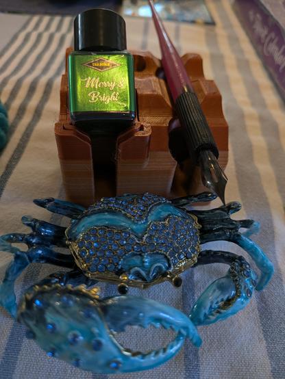 A blue enamelled crab ornament in front of a stand with a dip pen, with Diamine's Merry and Bright ink on it.