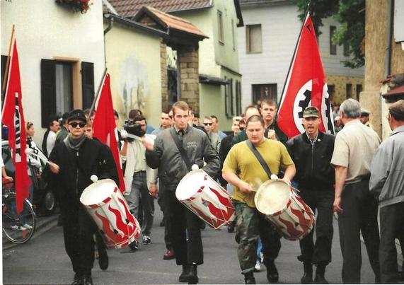 Neo-Nazi-Aufmarsch in Biebelsheim 1992: Drei Nazis trommeln, dahinter rote Fahnen mit einem weißen Kreis in der Mitte, in dem ein schwarzes Zahnrad ist — dort, wo früher mal das Hakenkreuz war.