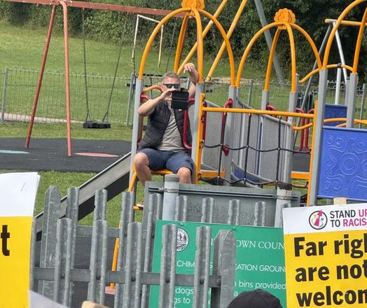 A man sits on a kids playground treehouse photographing marchers with his phone.
