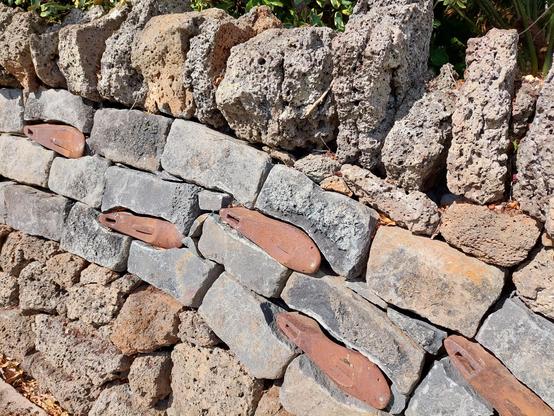 Wall made if volcanic stones at the bottom, then three rows of what might be bluestone above and then tipped by uneven, decorative volcanic rocks. Some reddish-brown fairly slender solid shapes are interspersed in the bluestone layer