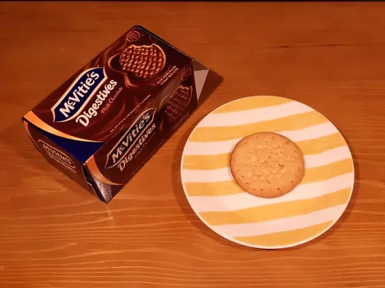 The image shows a light wooden table with a round plate on it. The plate has yellow and white stripes. On the plate, there is a single cookie that looks round and slightly textured. Next to the plate, there is a box of McVitie's Digestives. The box features a dark brown design and has a picture of the cookies on it. The cookies appear to be chocolate-covered. The setting looks simple and clean, with a focus on the cookie and the box.