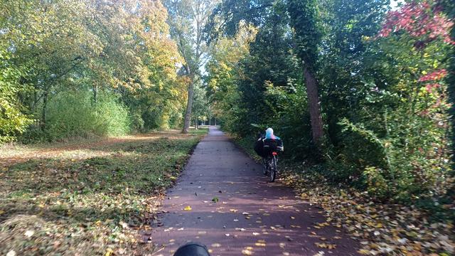 fietspad met bomen in herfstkleuren. voor mij rijdt een dame op een ligfiets met vrolijke pastelkleurige sjaaltjes in heur haar. topje van mijn linkervoet in beeld onderin