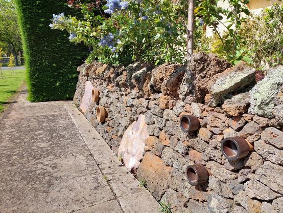 Drystone wall with brown clay pipes sticking out of it and a couple of large pale pink rocks among the smaller volcanic brown rocks.