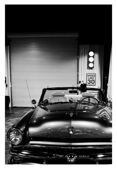 Black and white image of a classic convertible in a garage. A pair of dice hang from the rearview mirror, with a speed limit sign and garage door visible in the background.