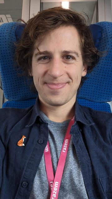 Moje zdjęcie selfie na niebieskim siedzeniu w pociągu. Uśmiecham się, mam na sobie granatową koszulę, szarą koszulkę, różową smycz z napisem Razem oraz przypinkę z liskiem.