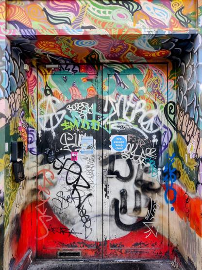 A doorway with layers of urban art and graffiti, a colourful mural of leaf like images, on top what looks like a faceless figure with black hat and red shirt, lots of tags and graffiti. A flyer and fire escape sign. Two doors, one with low letterbox.