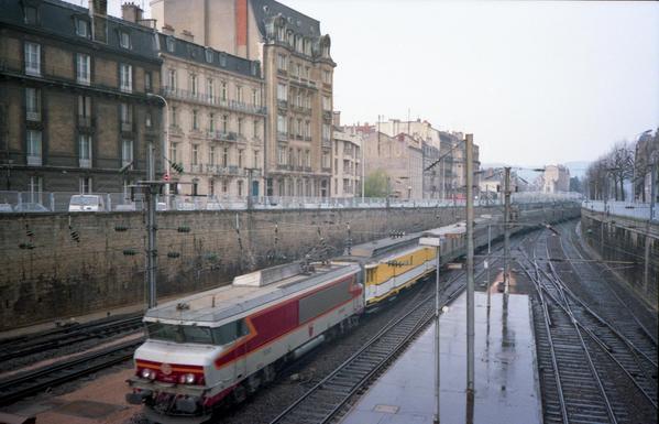 Voies de chemin de fer en tranché le long d'immeubles. Une locomotive rouge et orange tire un wagon jaune La Poste suivi de voitures passagers.