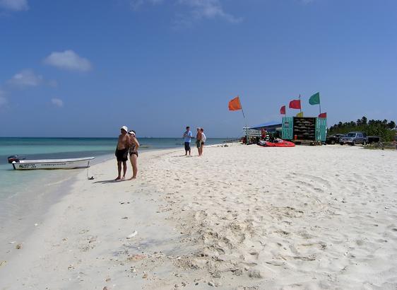 A few people are standing on a wide, white sandy beach looking out at the ocean. There is a small motorboat tied to the shore, and in the background is a small business that rents boards and other items. It is a small building painted aqua to match the color of the water. There are multiple, different colored flags flying above the building. Some cars and trucks are adjacent to the building as well as some tall bushes. The sky is blue with just a couple of clouds, and the ocean is very clear, going from pale aqua to deep blue in the distance. Aruba