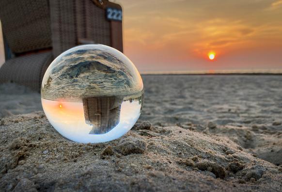 Foto mit einer Glaskugel. Am Sandstrand beim Sonnenuntergang. Die Kugel liegt vorne links der Mitte auf einem kleinen Sandhügelchen. In ihr spiegelt sich ein Strandkorb und der Sonnenuntergang. Durch die Kugelform steht alles auf dem Kopf, die Perspektive verzerrt sich, man sieht noch ein Stück blauen Himmel, der so nicht im Blickweld ist. 
Hinter der Kugel ist das Bild leicht unscharf. Es ist Ebbe, man sieht nichts von der Nordsee, nur Sandstrand. Der Strandkorb selbst ist im Foto nur zum Teil sichtbar. Er steht am linken Rand, der obere Teil ist angeschnitten. Die Sonne, knapp über dem Horizont, leuchtet rechts der Mitte und taucht den Himmel in Orangetöne.
